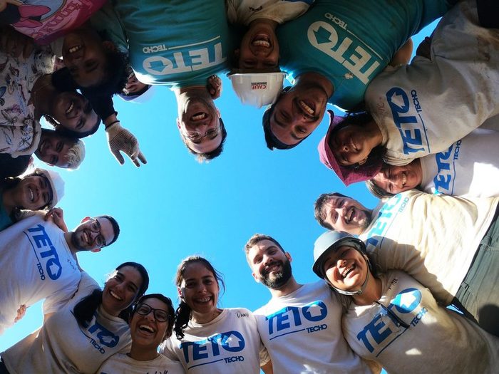 Voluntários da TETO em roda, vistos de baixo, sorrindo contra o céu azul