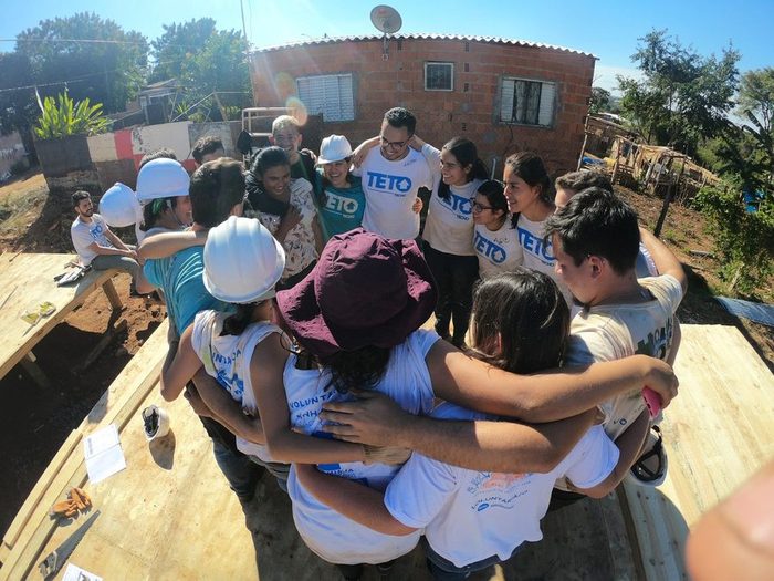 Equipe de voluntários abraçada em roda sobre o assoalho da casa recém-construída